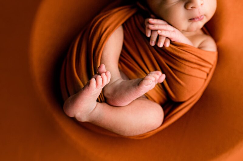 Close the image of a baby's feet is his wrapped in a rust colored wrap during his Studio newborn session