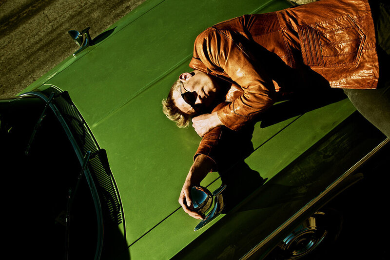 Musician photo Ryan Guldemon lying on hood of green vintage car wearing orange coat and sunglasses Route 66