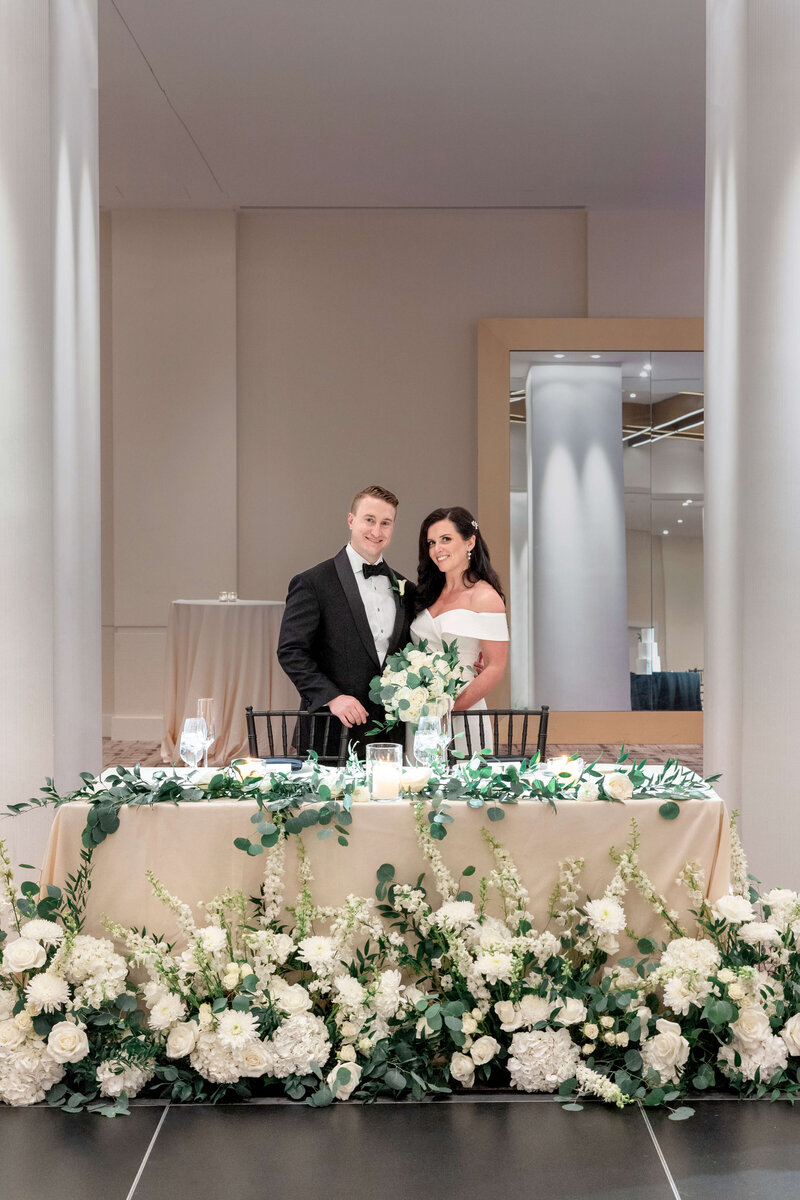 A couple stands behind a table adorned with white flowers and greenery. The man wears a black tuxedo, and the woman wears an off-the-shoulder white dress, holding a bouquet. The setting is elegant, with candles and a mirrored wall in the background.