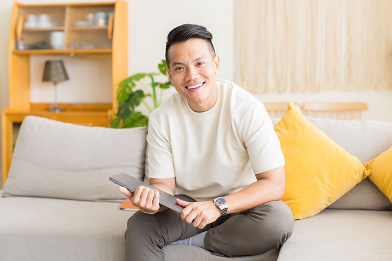 Sho Dewan, young Asian man, career coach and founder of Workhap, sitting on a light grey couch with yellow cushion, with his laptop in hands, looking and smiling directly at the camera