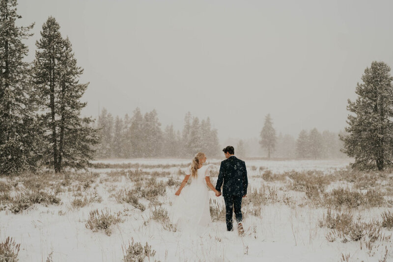 Man and woman walking away from the camera, holding hands in a snowy outdoor forest