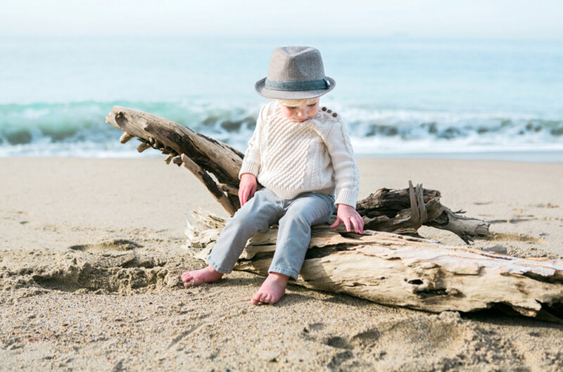 A little boy wearing a gray hat sits on a piece of driftwood on the beach