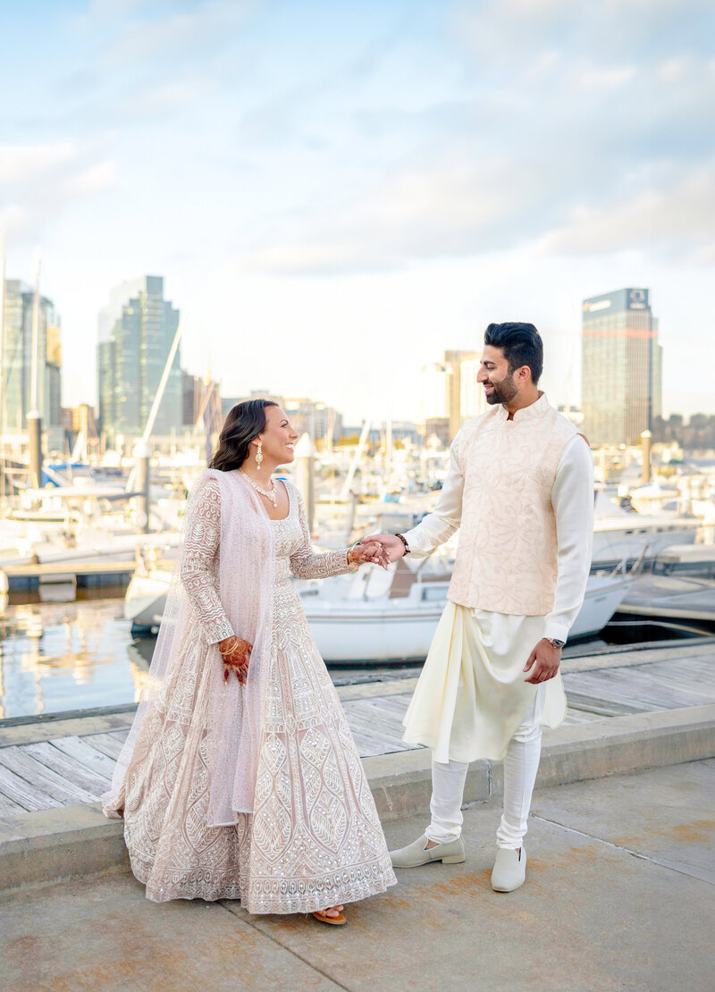 A couple in elegant traditional attire smiles and holds hands on a dock with boats in the background. The woman wears a detailed white dress, and the man is in a cream ensemble. City buildings are visible in the distance under a partly cloudy sky.