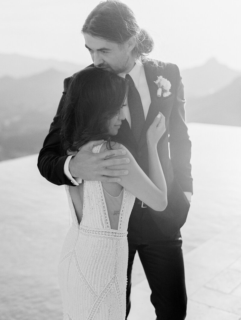 black and white photo of couple hugging with Malibu hills behind them. She has a beaded low back wedding dress on. He has a slim fitted suit and black tie