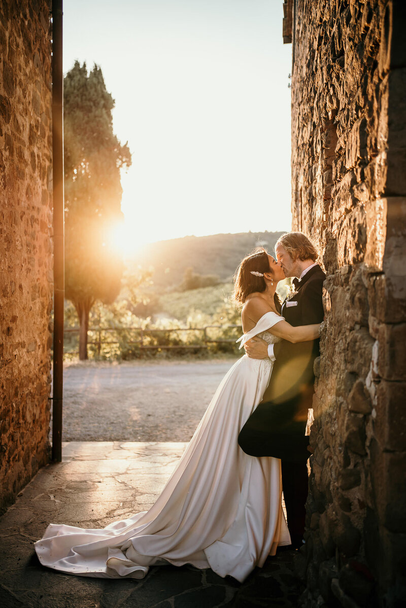Groom leaning on a stone wall while bride is kissing him during the sunset in Chianti
