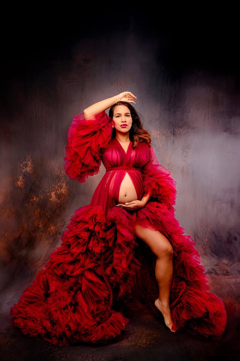 Expectant Charlotte mom stands in a dramatic pose and a red robe with tons of layers one hand above her head the other on her belly leg extended on a purple background