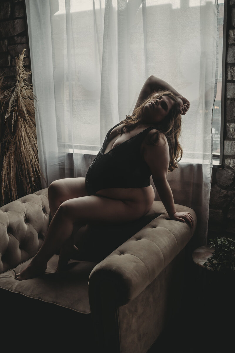 Natural light boudoir portrait of a curvy woman posed on a chair with moody natural light