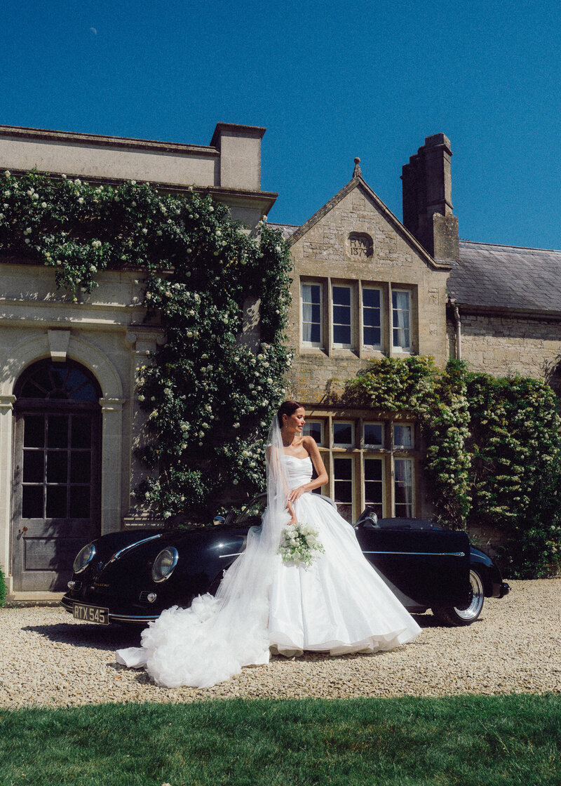 Bride standing beside a classic black wedding car in front of an elegant historic estate, captured in a romantic, cinematic style by a luxury documentary wedding photographer UK.