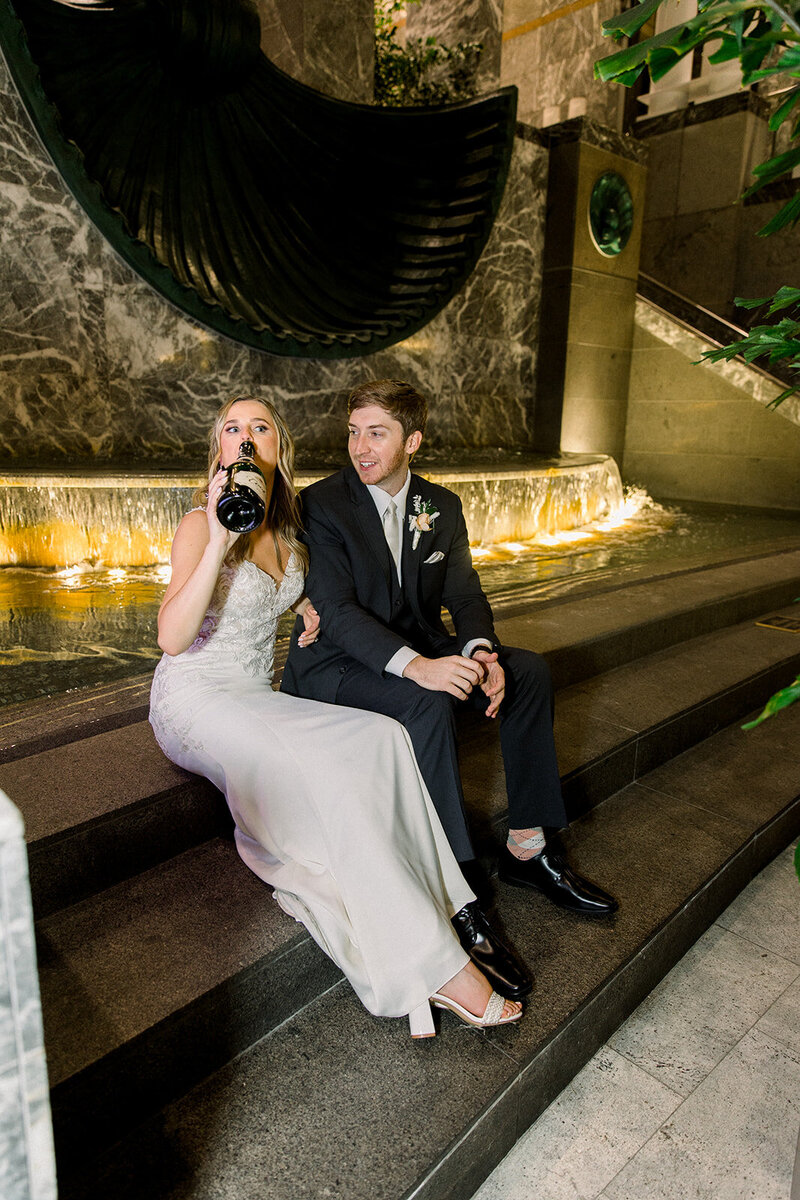bride drinking champagne from bottle near fountain