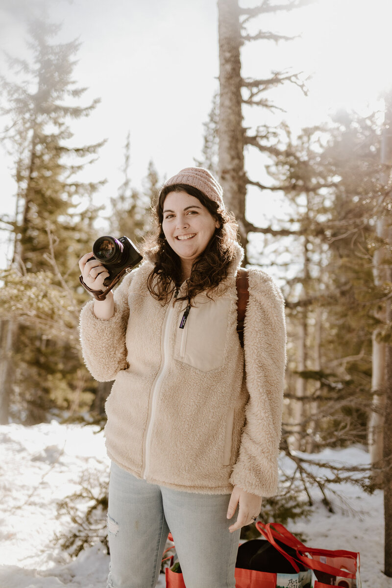 Mt Hood Elopement Photographer Lindsey Wickert stands in a snowy forest in a beige sweater in a snowy forest holding her camera