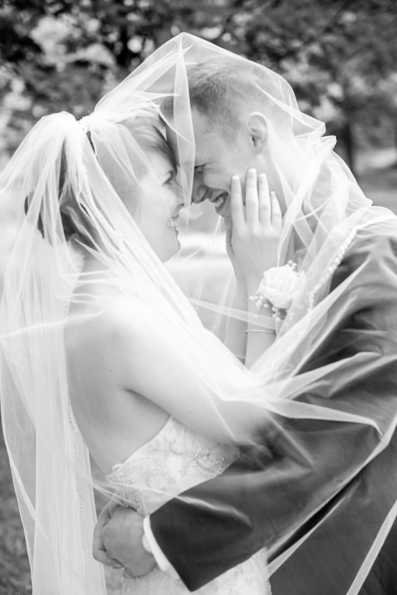 A black and white photo of a bride and groom underneath her wedding veil.