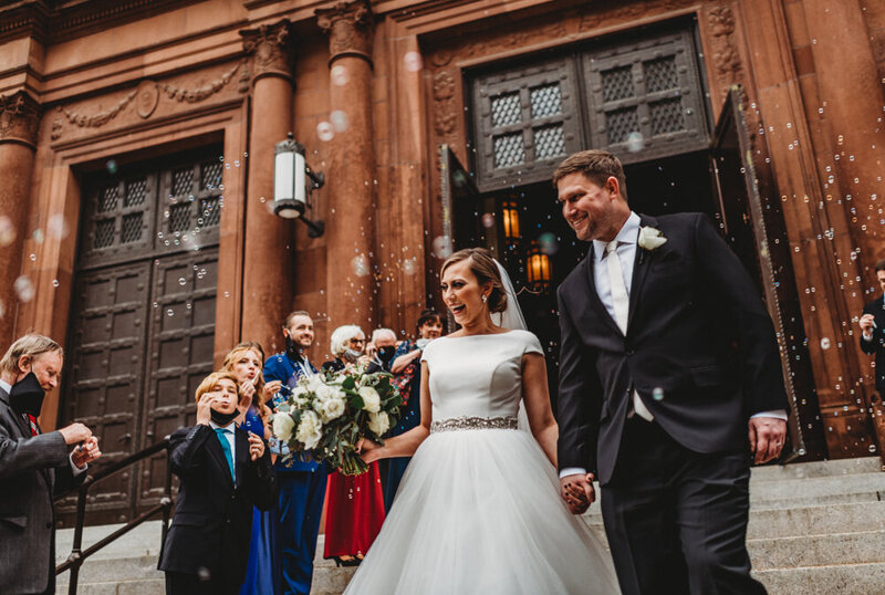 Wedding day exit captured by Maryland wedding photographer with bride and groom holding hands as they do send down the stairs from the chapel while their guests blow bubbles and cheer for them