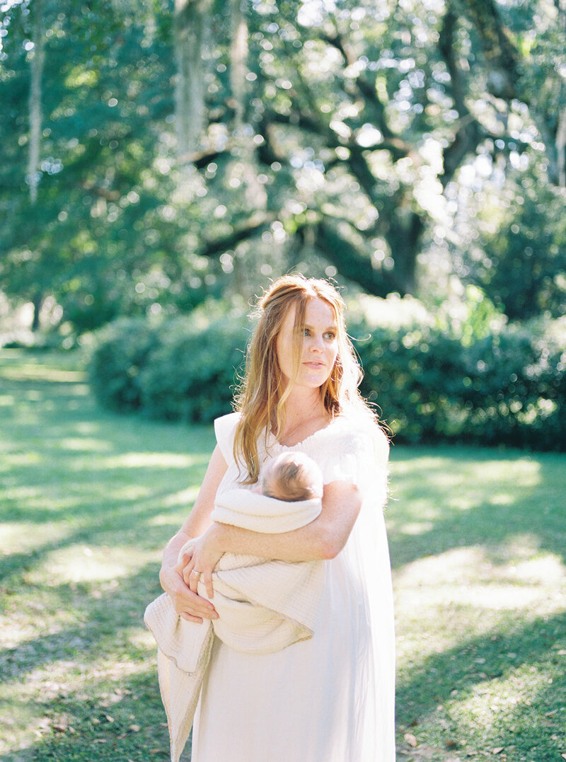 A new mother stands in a green pasture,  holding her newborn swaddled in a white blanket while looking off into the distance during her newborn session