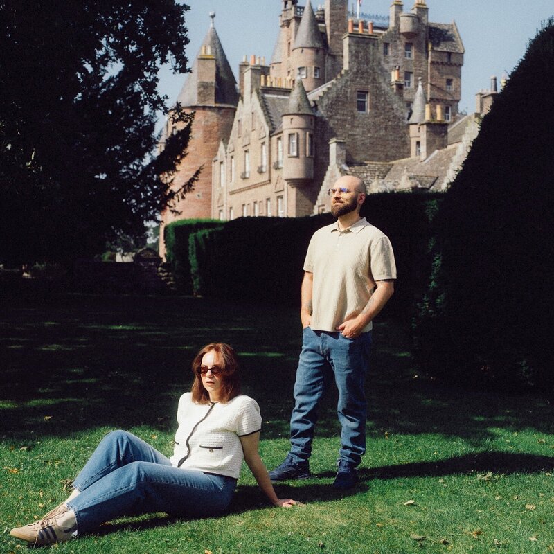 A husband and wife photography duo pose on the lawn of a historic castle, one seated and the other standing, creating a relaxed and confident scene.