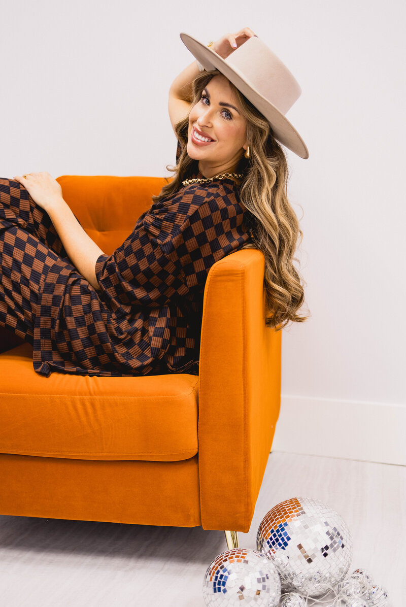 woman wearing a wide brimmed hat sitting in an orange chair while smiling