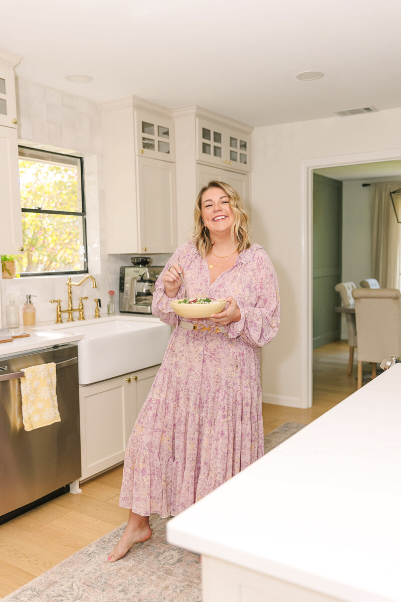 Mollie Mason in a purple dress holding a bowl of food and smiling while standing in her kitchen