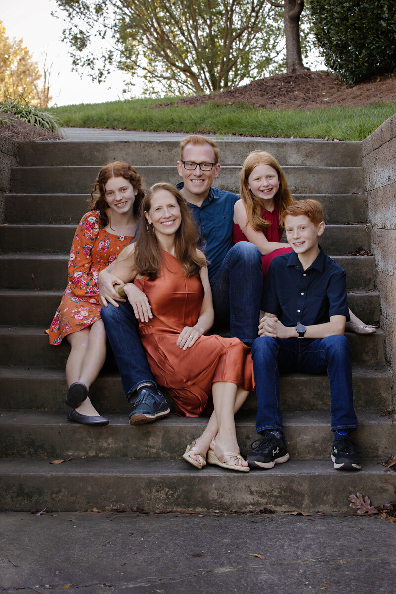 Charlotte photographer captured enchanting photo of a family of five posed together on a concrete staircase. Family outfits include Mom in a beautiful orange long silk dress, daughter in an orange floral dress, son in a blue short sleeve button down with jeans and Dad in a similar outfit and youngest daughter in a red dress.