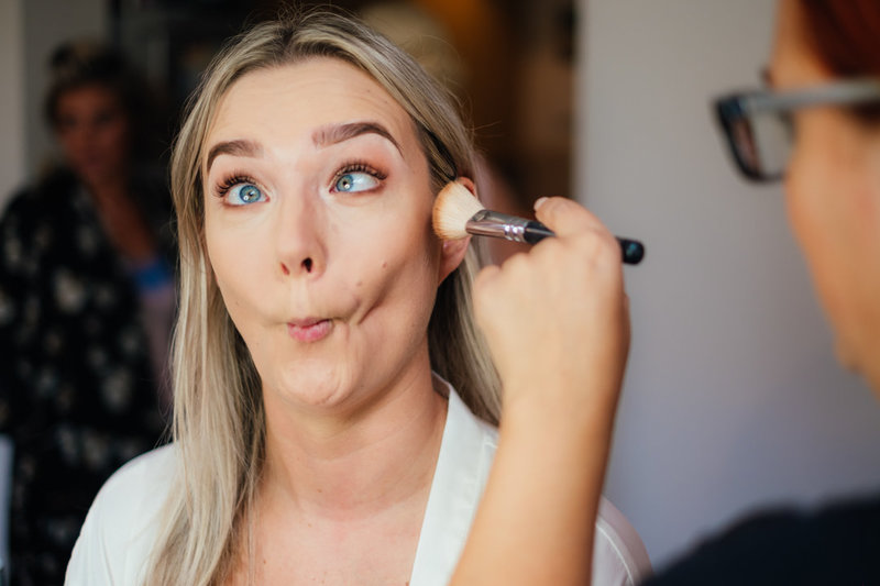 bride sucks her cheeks in and makes a silly face as she has her makleup applied the morning of her wedding day