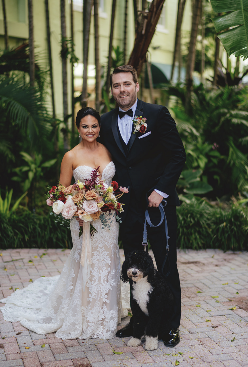 short tan bride wearing strapless, lacy gown hold large, colorful bouquet next to groom holding black and white dog on a leash.
