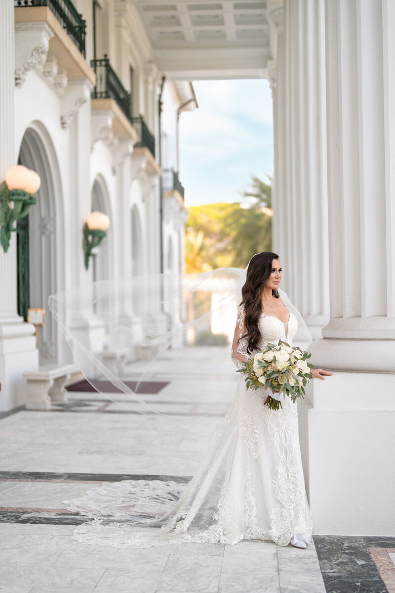 A bride in a lace gown holds a bouquet and stands beside grand white columns, her veil flowing behind her, symbolizing elegance and luxury wedding planning.
