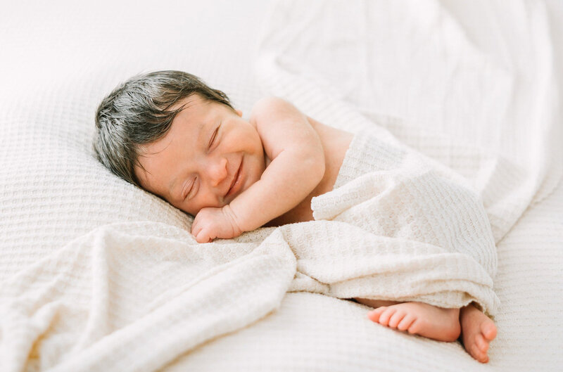 A newborn baby girl with a full head of dark hair smiles in her sleep during her newborn session with Daniele Rose Photography