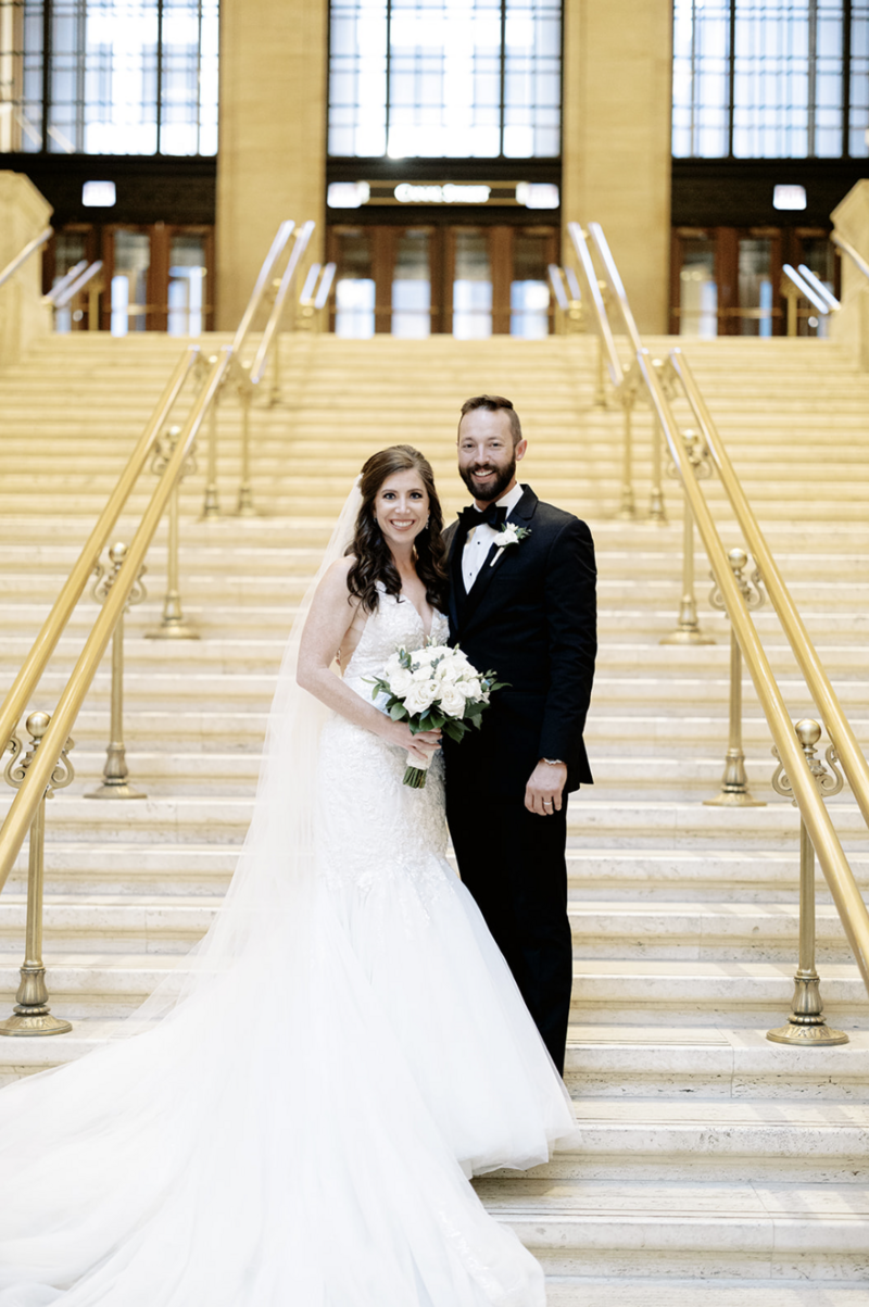 Bride with brown hair holds bouquet and smiles with groom in black tux in front of a staircase.