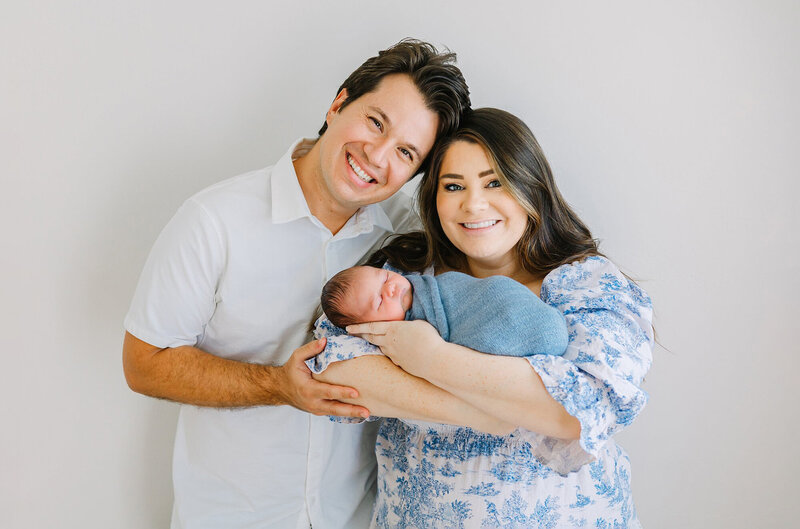 A family smiles while holding their newborn baby boy who is wrapped in a blue swaddle in Daniele Rose photogrpahy's Camarillo newborn studio