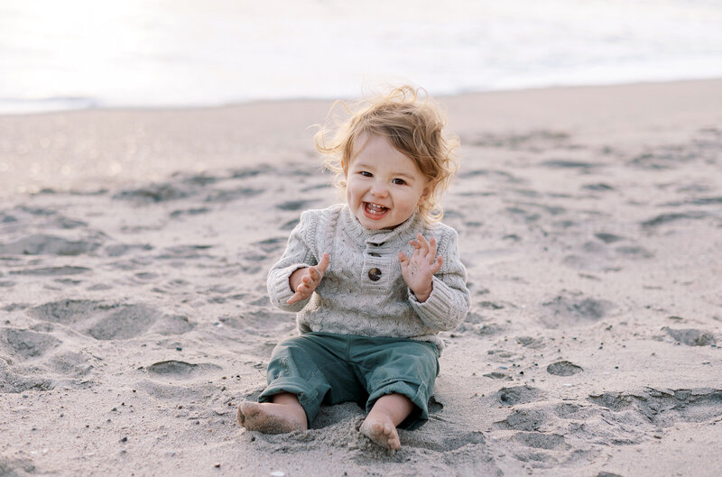 A little boy in a chunky knit sweater laughs and smiles at the camera at Will Rogers Beach