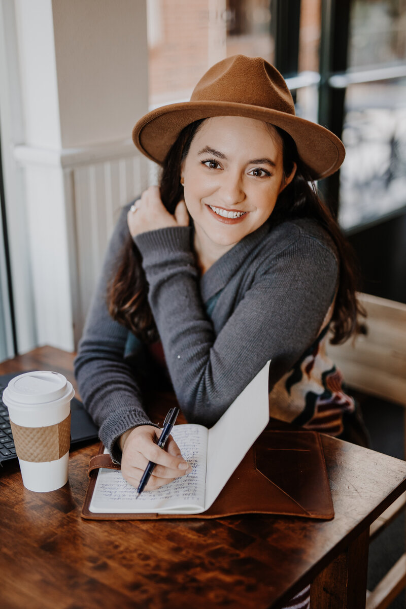 Orlando Wedding Photographer Joanna of Four Loves Photo and Film is sitting at a coffee shop, smiling at the camera with a pen in her hand, writing in a journal, with a cup of coffee on the table next to her.