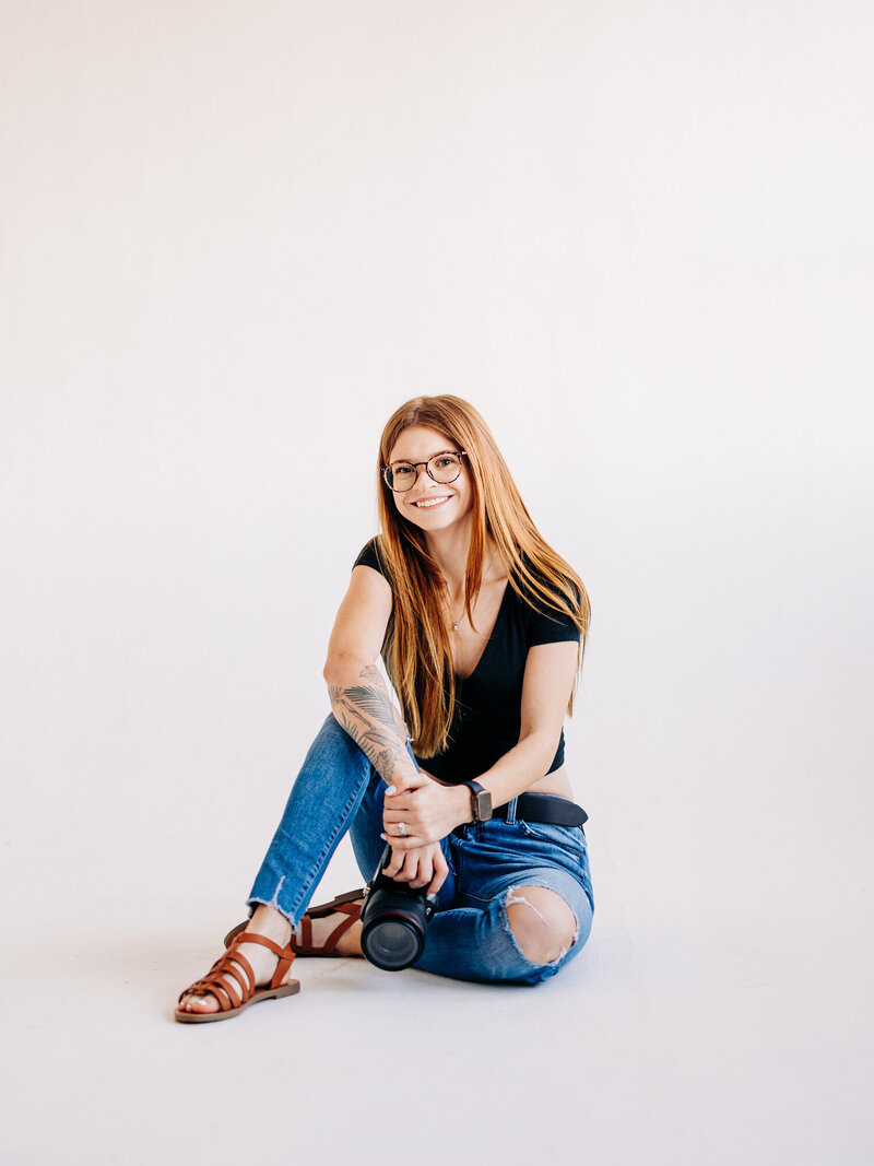 This image features San Antonio Wedding Photographer Kylie Jacobsen, owner of KD Captures, sitting on a stool. The image was taken at Collective Studios in San Antonio, TX. She is holding a macbook pro in her lap, and wearing a black shirt and grey plaid pants.