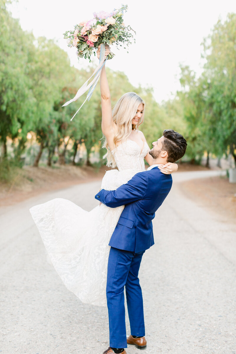 A bride pointing her bouquet up the sky while the groom lifts her up at Eden Gardens in Moorpark California.