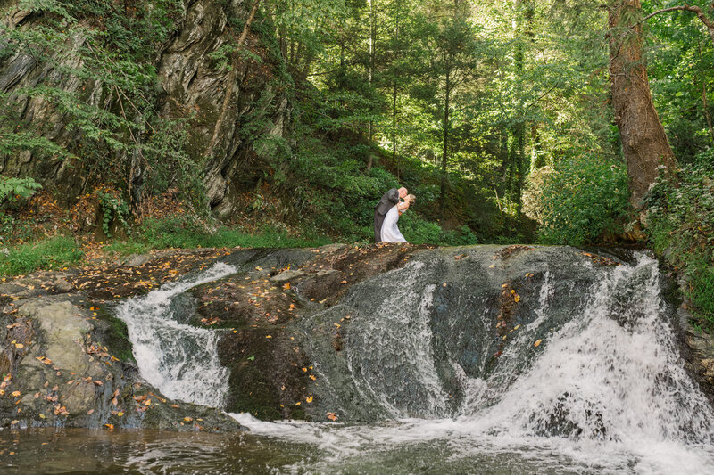 A bride and groom kissing while standing at the top of a small waterfall.