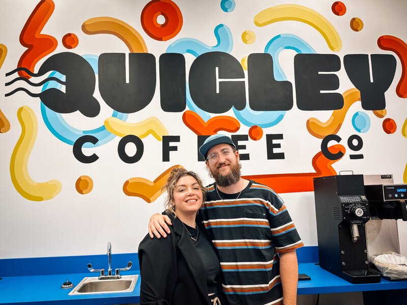 Quigley Coffee Co founders standing in front of colorful mural logo, representing the brand story and leadership behind the franchise.