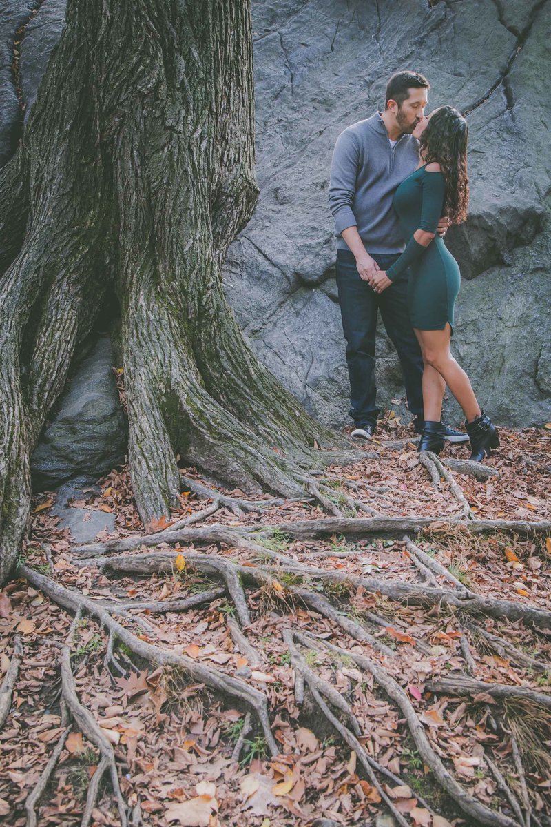 A couple kisses in Central Park near tree roots during their engagement session.