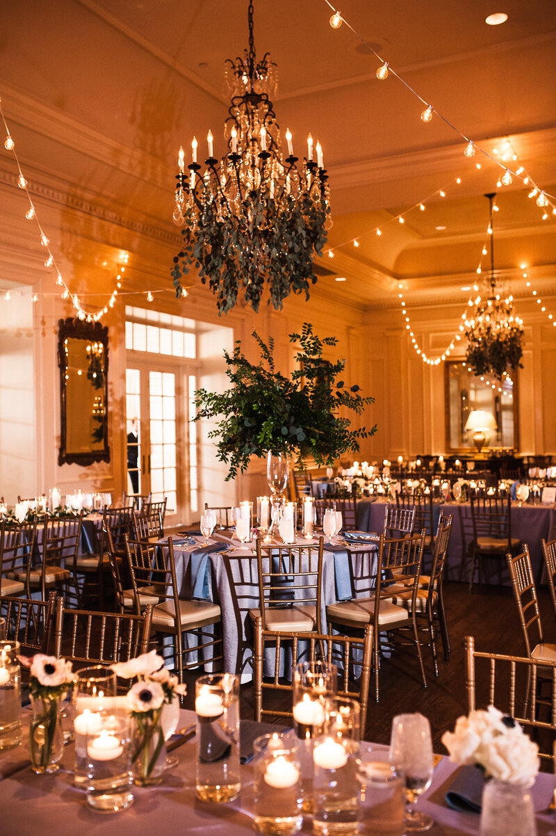 Wedding reception at country club ballroom filled with elegant tables adorned with white candles and flowers. Ballroom ceiling draped in soft twinkle lights and suspended greenery.