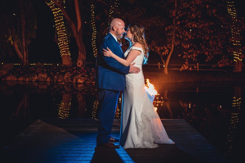 Bride and groom in an embrace on the dock of the lake with trees and lights at Lake Oak Meadows wedding venue in Temecula.
