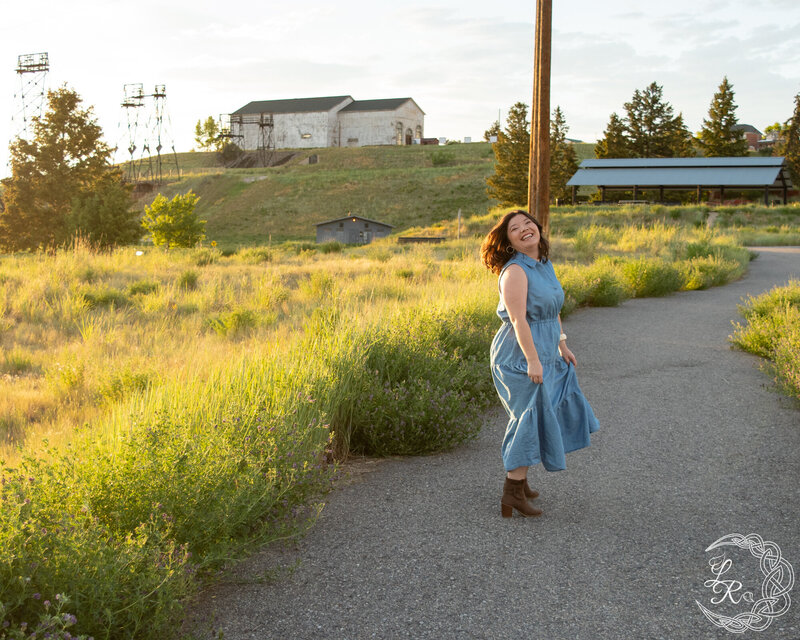 photo of Ashlyn standing on a sidewalk in a blue denim dress, tossing her hair over her shoulder and smiling. alfalfa, grass and headframes int he background