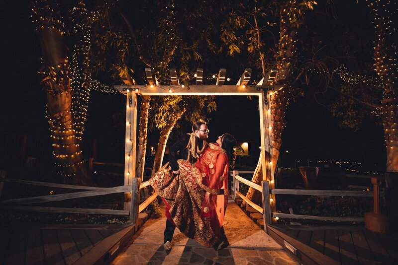 Bride and groom in a dip wearing Indian wedding attire under an arch with twinkle lights and trees at Lake Oak Meadows in Temecula.