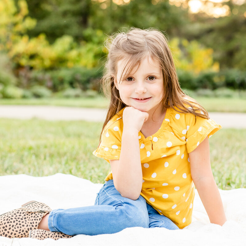 Little girl with yellow shirt sitting with chin in hand outdoors on a blanket for family session