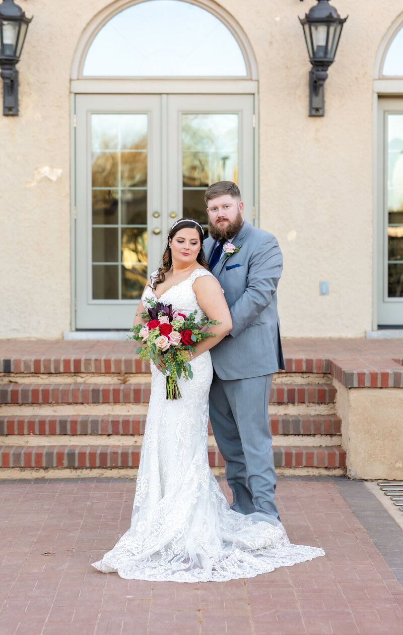 Bride is wearing a white lace sleeveless dress holding a bouquet and looking down her shoulder while groom is wearing a gray suit and looking at the camera.  The couple are standing outside at the Texas Tech Merket Alumni Center