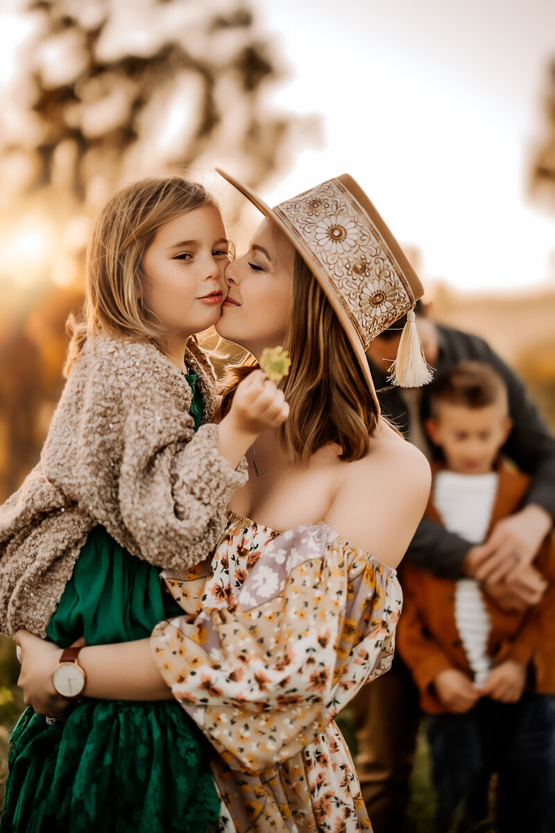 Family Photographer,  Mom wearing a floral dress kissing her daughter's cheek with dad and son in background