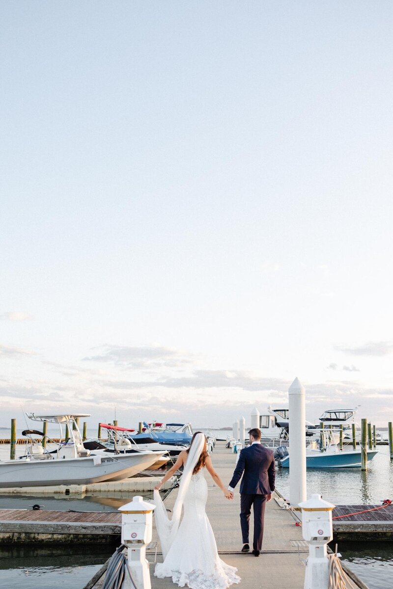 Newlyweds walking hand in hand on a dock with boats in the background, captured by a luxury wedding photographer.