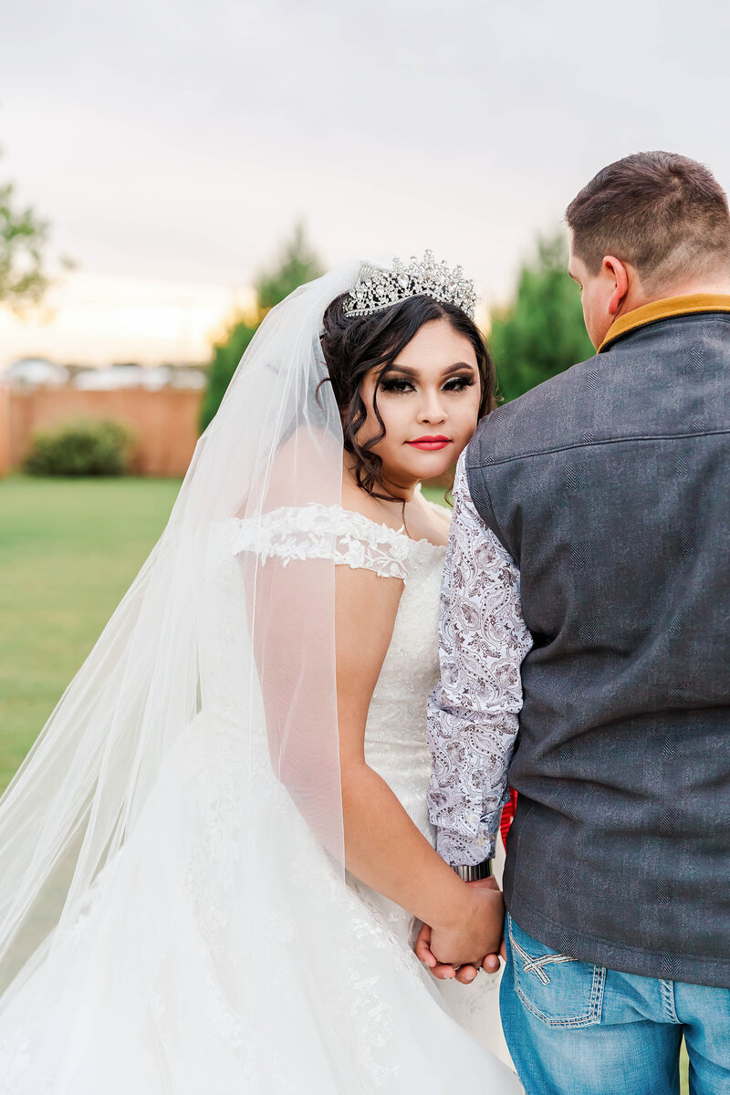 Hispanic bride looking at camera wearing a white  sleeveless wedding dress with a long veil while groom is looking at the bride