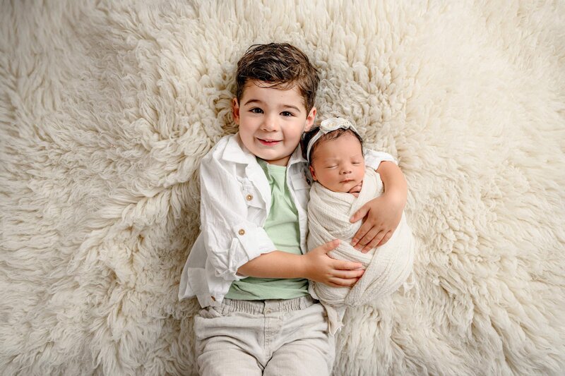 toddler boy dressed in green and neutral colors holds his newborn sister wrapped in cream on a cream rug