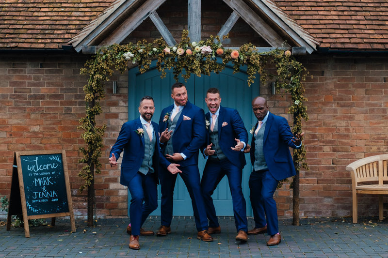 5 groomsmen dressed in blue suits with tan shoes make silly poses, faces and gestures in front of a pretty flower decorated wooden doorway at their swallows nest barn wedding