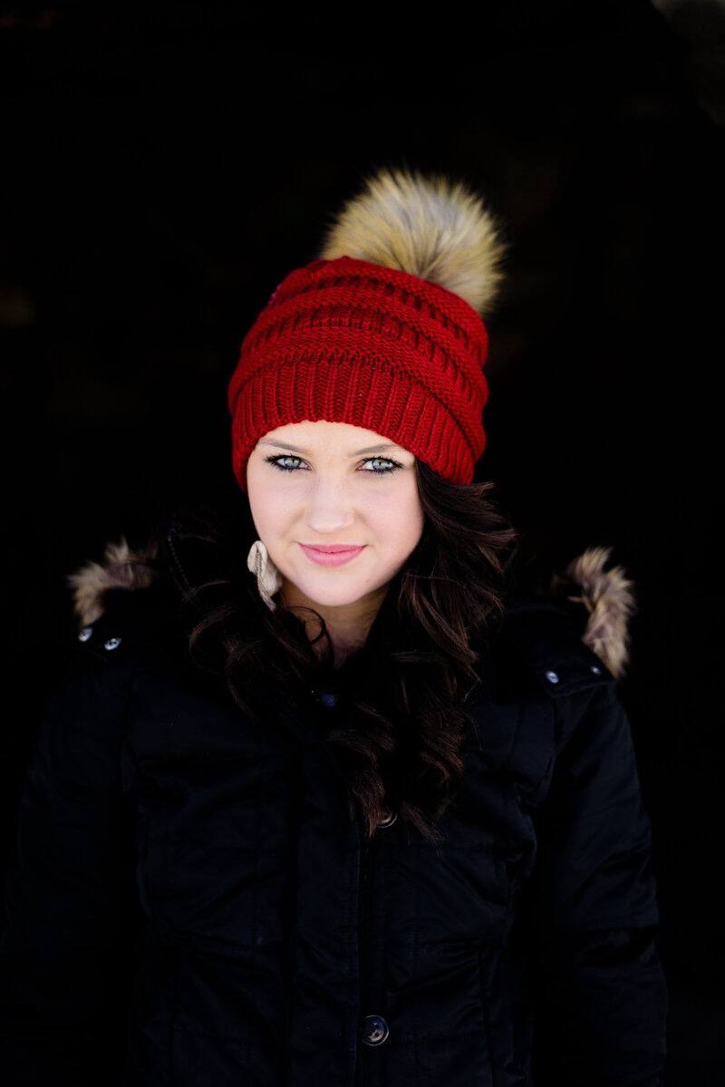 Senior outfit ideas of a girl with bright green eyes wearing a red winter hat with a puff ball and a black fur lined jacket and a dark black background