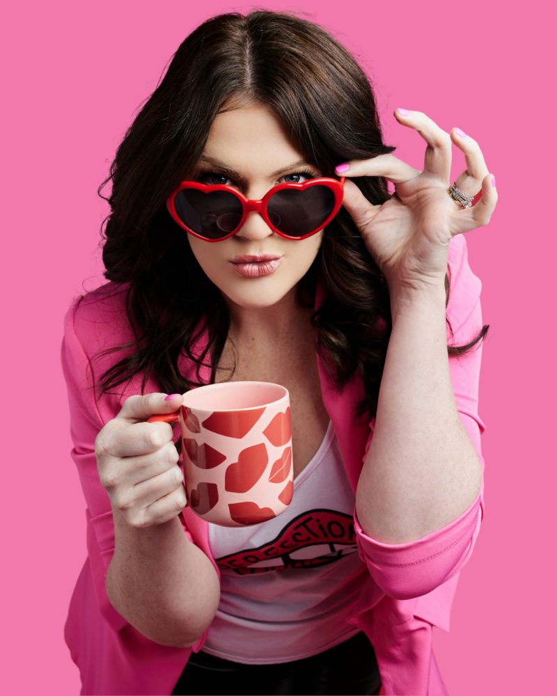 Emily wearing heart shaped sunglasses and holding a mug with lips all over it. She is pulling on the side of her glasses so you can see her eyes.