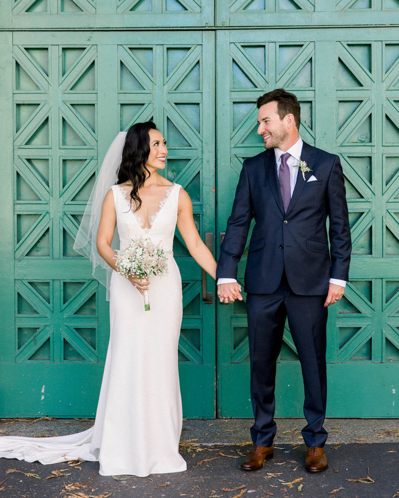 Bride + Groom standing looking at each other smiling holding hands in front of teal doors