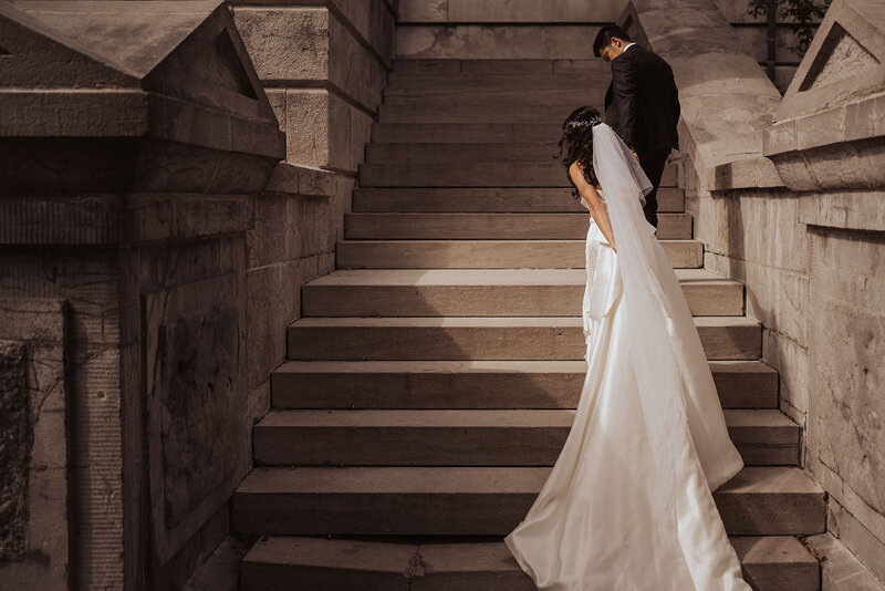 Photograph of a bride ascending stairs with her gown trailing, and the groom ahead.