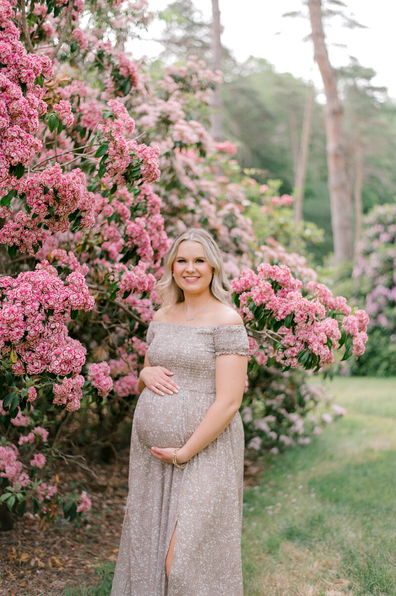 An expecting mother standing in front of a tall flowering pink bush as she holds her bump and smiles at the camera